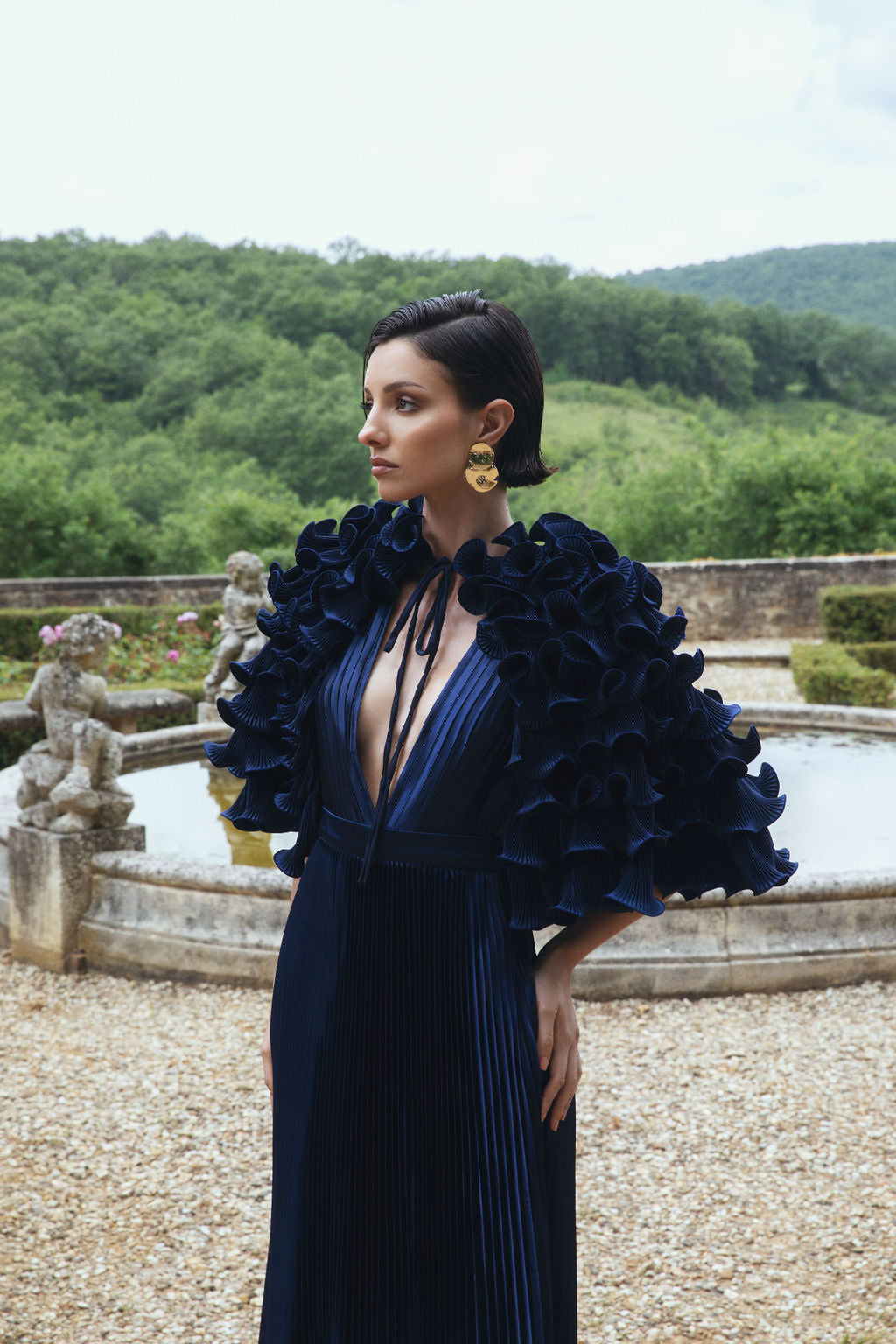 Model in a navy pleated dress with ruffled sleeves, standing by a fountain with a scenic landscape in the background.