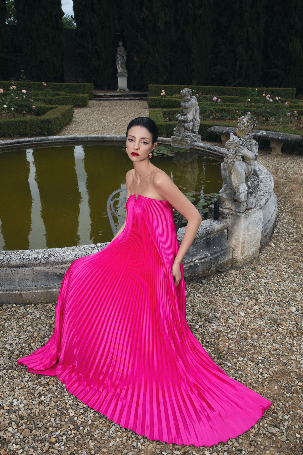 A woman in a pink dress poses charmingly before a fountain