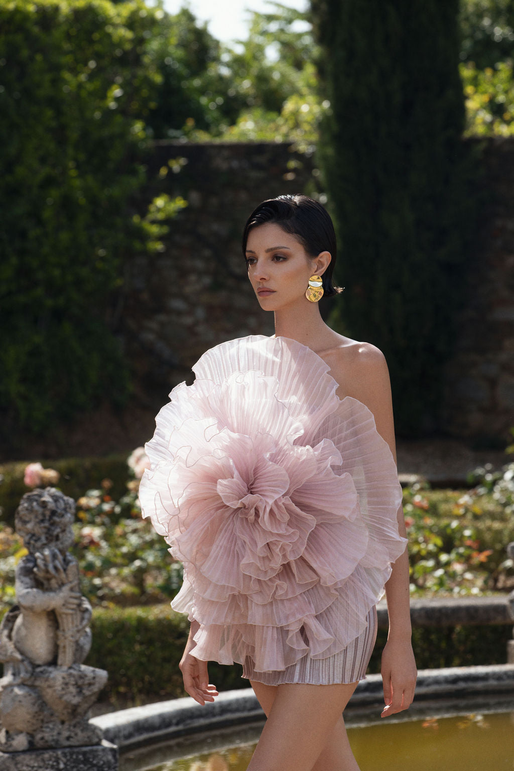 Model in a pink ruffled one-shoulder dress standing by a fountain.