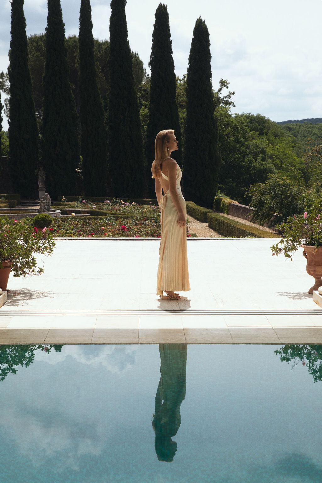 Model in a cream pleated dress standing by a pool in a formal garden