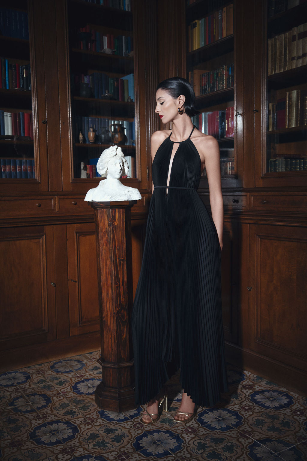 A woman in a stylish black dress stands before a bookcase